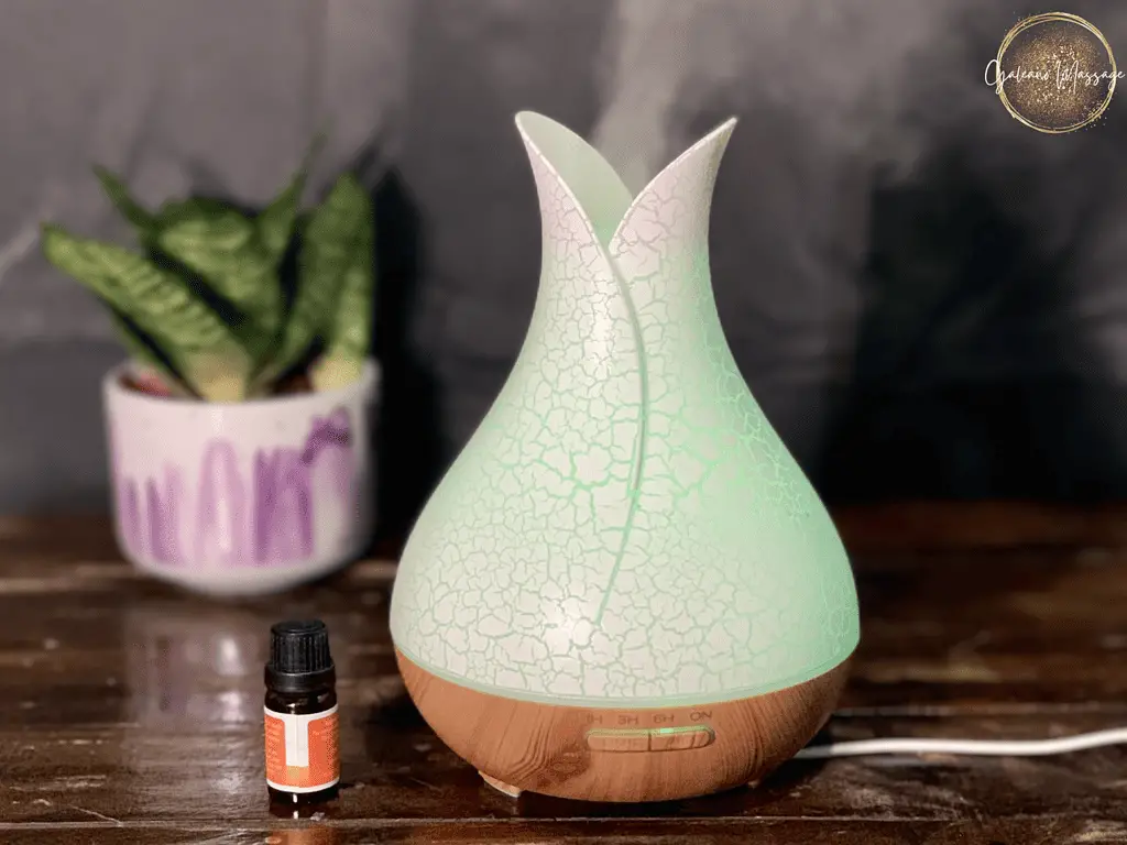 An aromatherapy oil diffuser with green light therapy is sitting on a wood table with a green house plant. Blurry dark gray curtain background. Gold and white Galeano Massage logo.